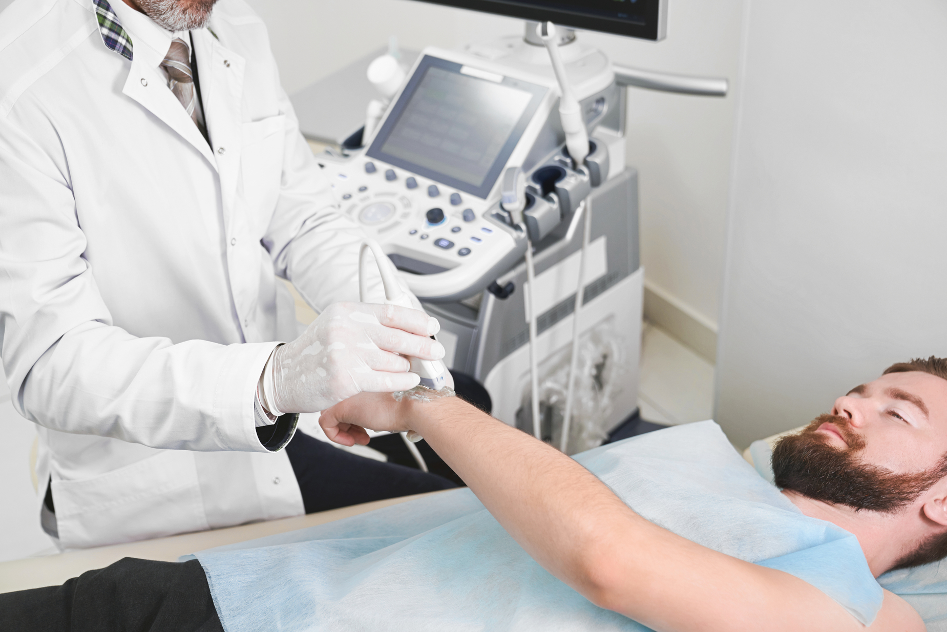 Bearded male patient lying in medical office, on ultrasound diagnostics. Professional therapist holding wrist of man, doing examination, using ultrasound probe and special equipment.